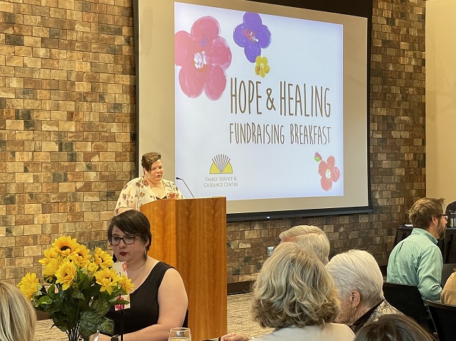 Female FSGC Staff Member At Podium Addresses Audience Seated At Tables. Projector Screen Behind Her Reads Hope And Healing Fundraising Breakfast
