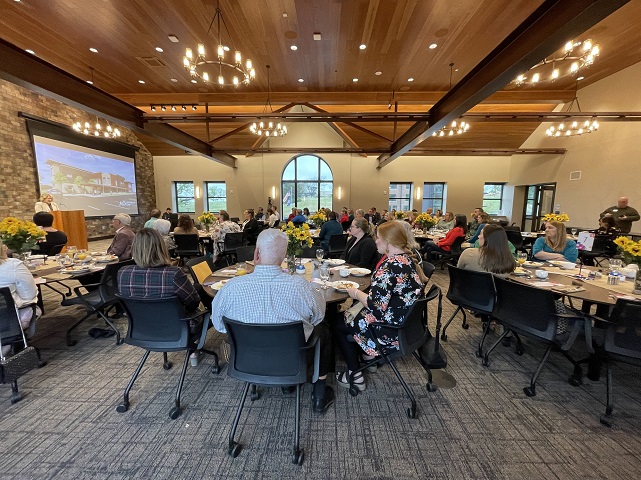 Photo Taken From Side of Room, Multiple Tables of Seated Guests Eating Breakfast at FSGC Hope & Healing Fundraising Breakfast. Female FSGC Staff Member Stands at Podium and is Speaking to Group. 