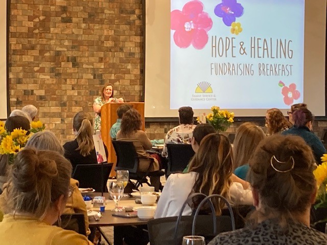 Jennie, an FSGC Staff Member, Stands At Podium and Speaks to People Seated At Tables During Hope & Healing Fundraising Breakfast