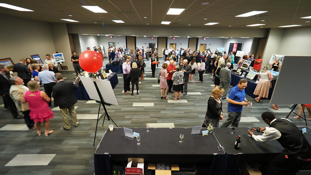 A wide overhead shot. The space is in a conference center, and various tables are set up displaying silent auction items. Crowds of people walk freely between the tables, and some are talking in groups in the middle of the room.