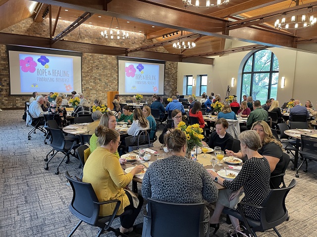 Wide Shot From Back of Room During Hope And Healing Fundraising Breakfast. Guests Are Seated at Round Tables with Sunflower Centerpieces. Two Project Screens At Front of Room Read Hope and Healing Fundraising Breakfast.