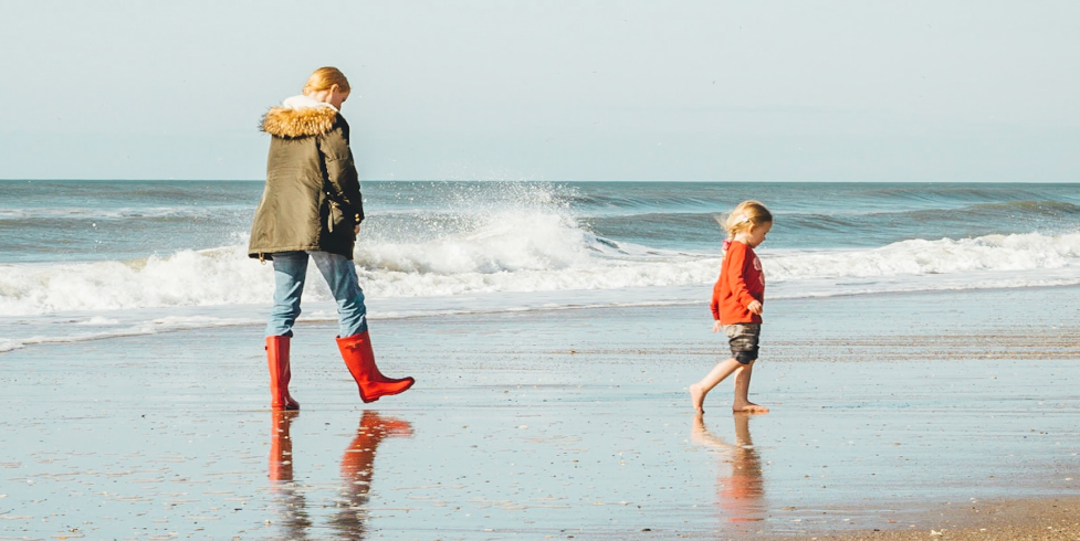 A woman follows her small child as they walk along the waves of a beach. The woman wears big red boots that reflect off the wet sand and the child's long hair blows in the wind