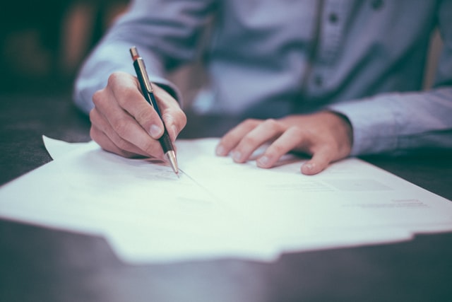 A man examines papers in front of him and uses a pen to sign. Only his torso is in frame.