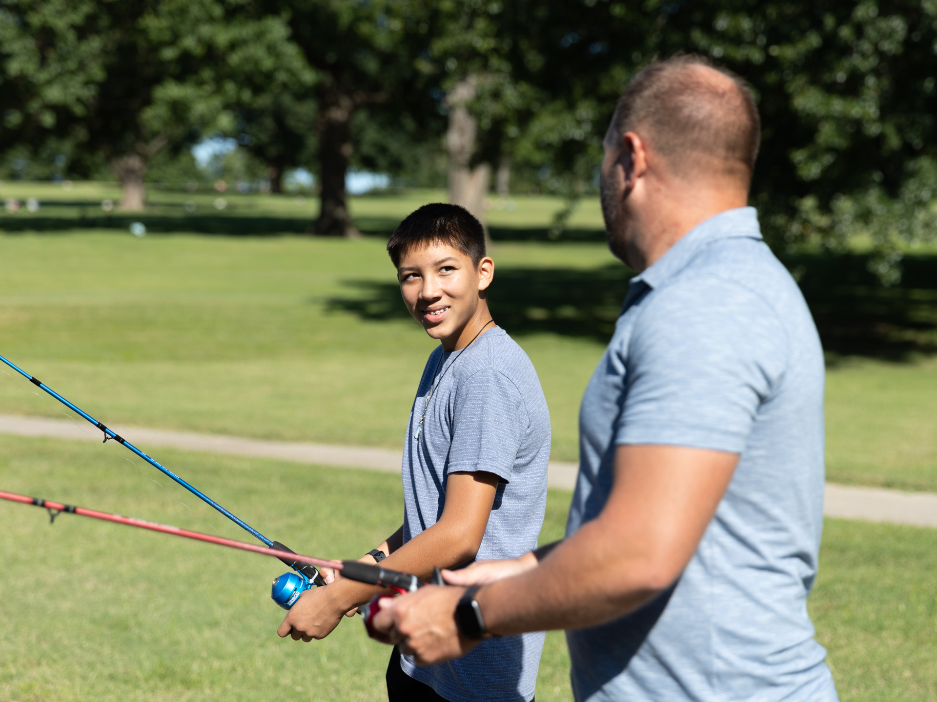 A man in a polo shirt stands along side a smiling boy as they reel in fishing rods at the park