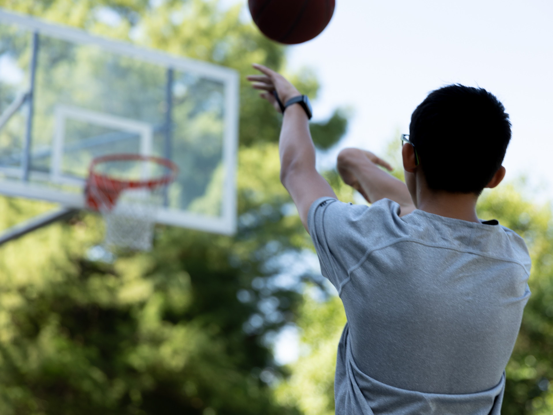 A young man faces a basketball hoop as he attempts to put the ball in the net. His arms are outstretched as the ball flys through the air.