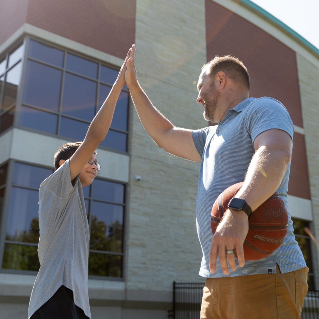 a man holding a basketball at his side highfives a young boy on the basketball court with a building behind them