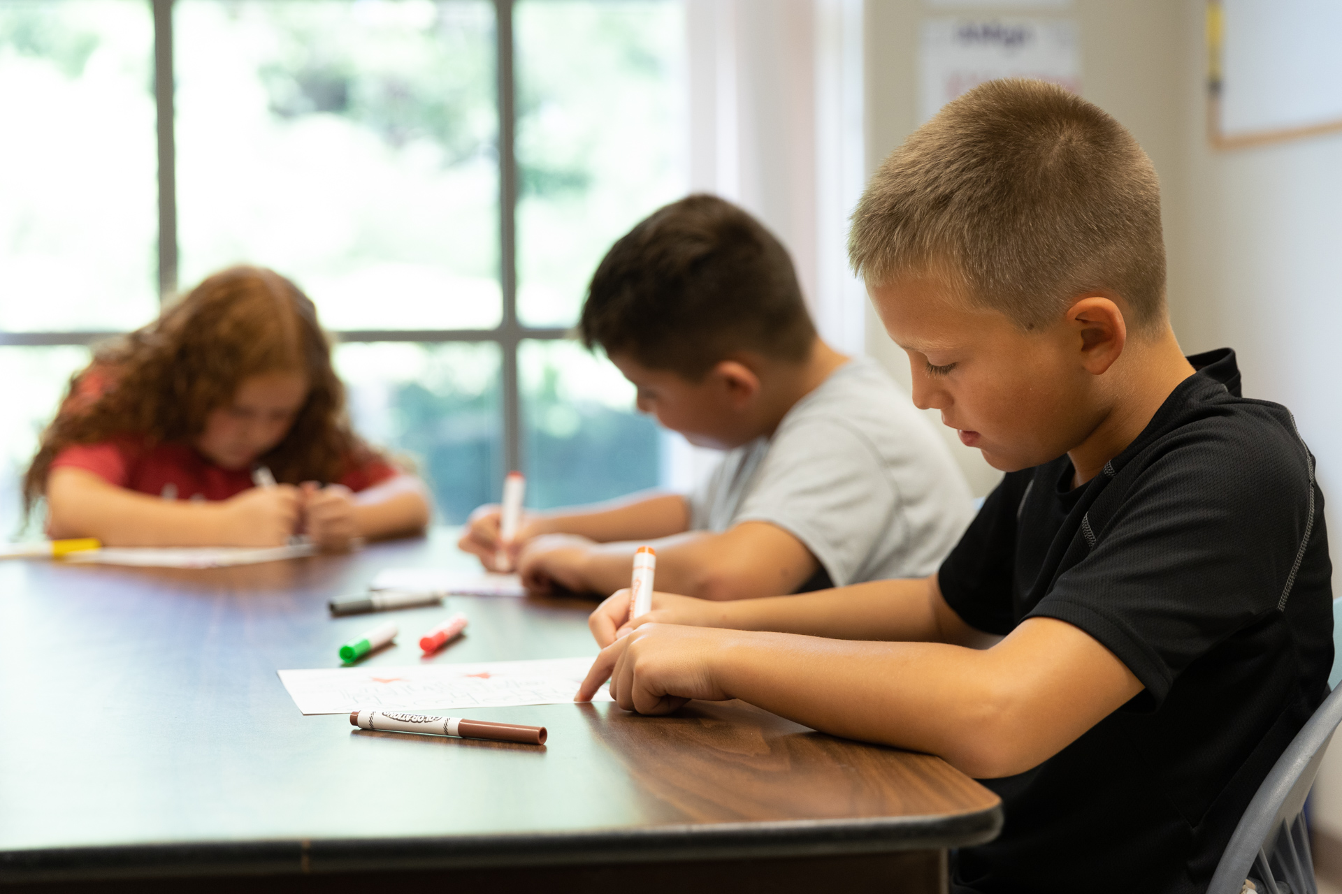 3 children sit at a brown table using various markers to draw their own pictures inside FSGC.