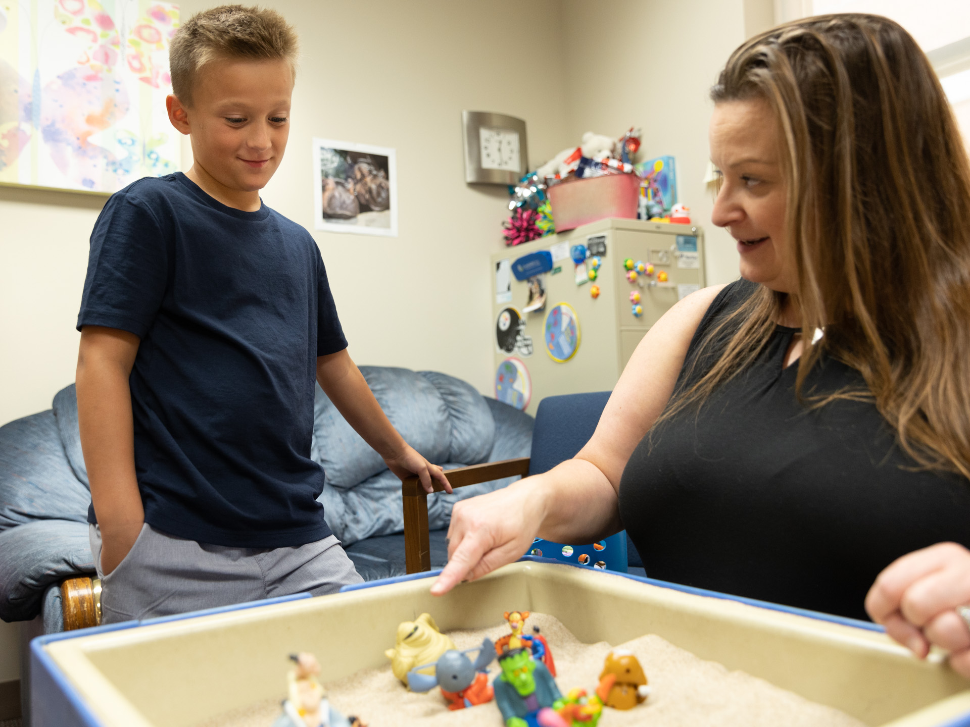 A talking woman points to a toy inside an indoor sandbox to a grinning young boy.
