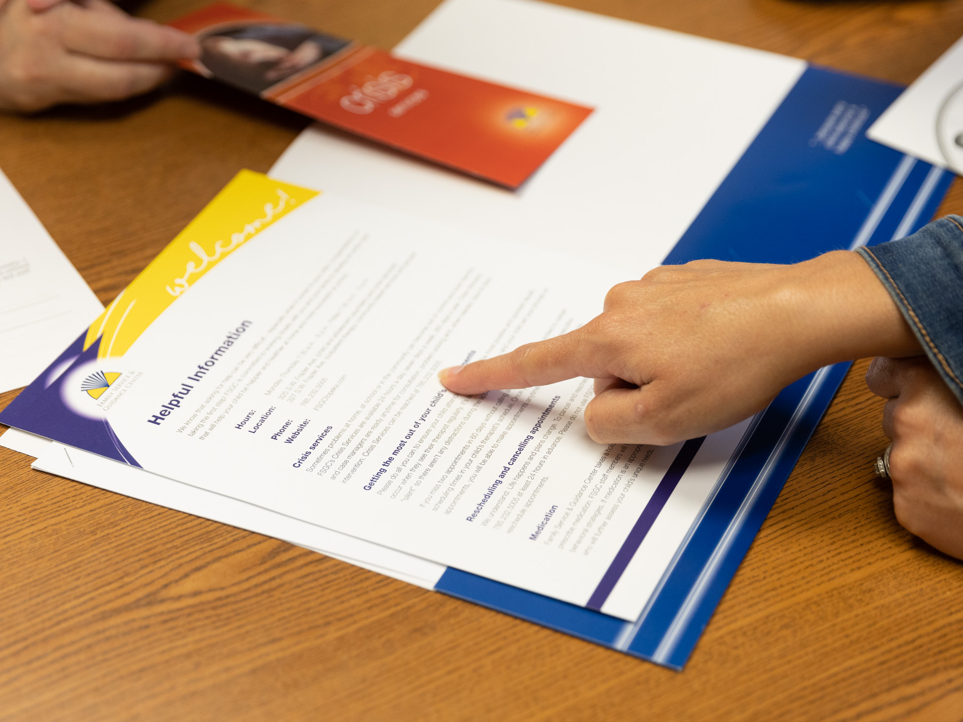 A finger points to a FSGC pamphlet that reads "Helpful Information", that lays on a wooden desk, an FSGC employee's hand can be seen on the other side.
