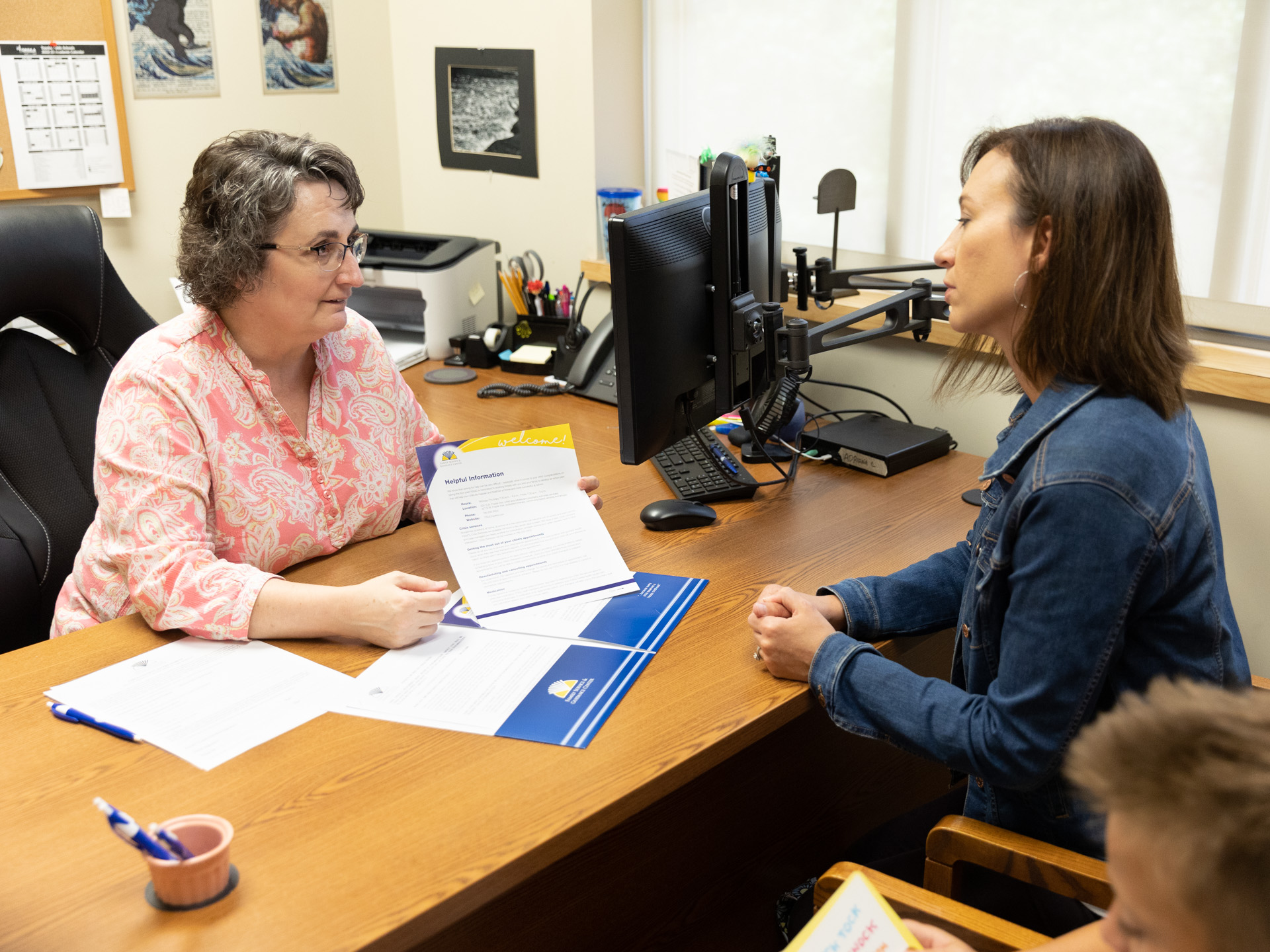 FSGC member, Pam Evans, sits at an office desk across another woman, showing her a "helpful resources" brochure.