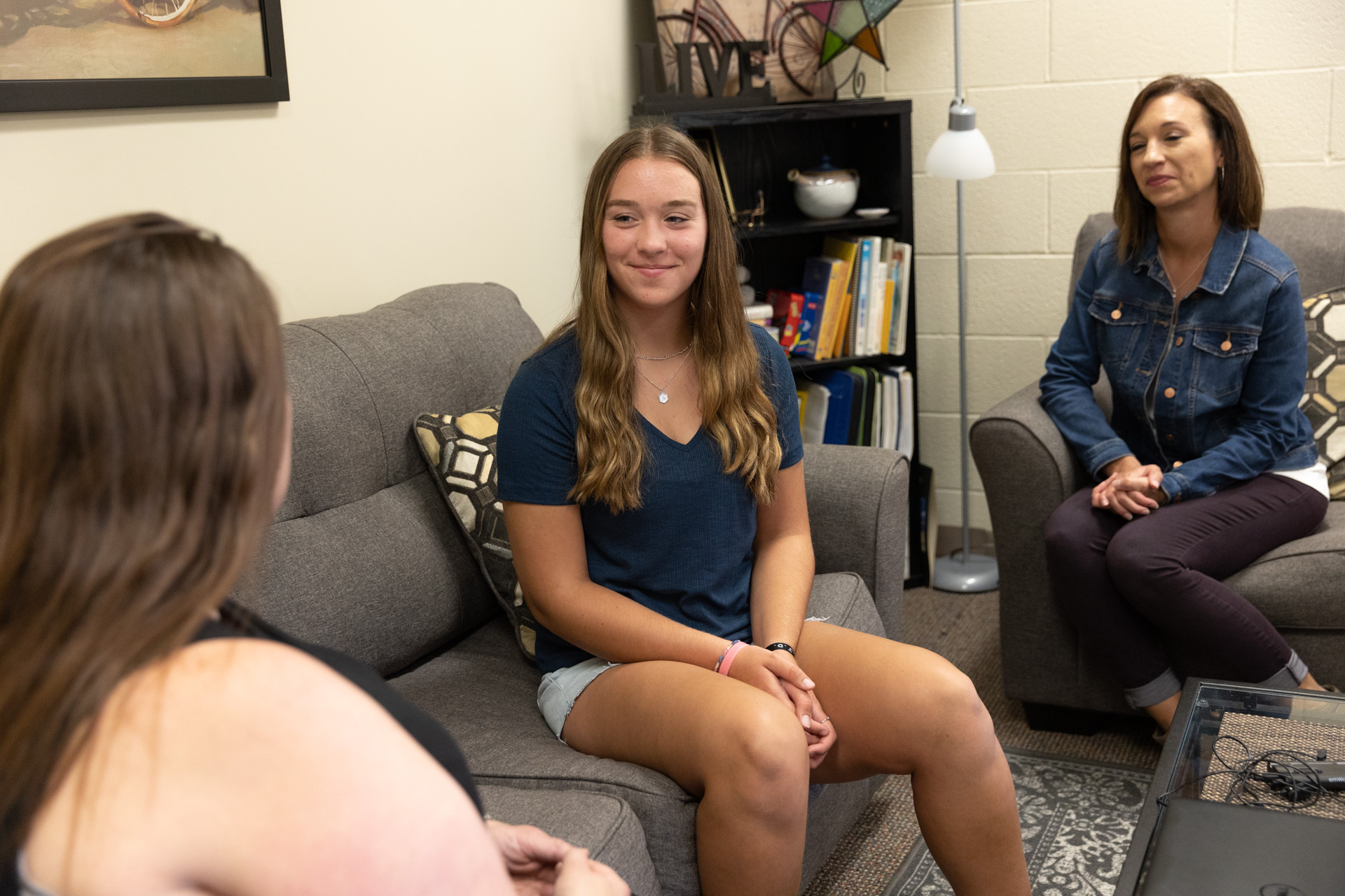 A smiling young girl sits on a couch facing a FSGC worker. Behind the young girl, a woman sits with her hands folded on a different couch