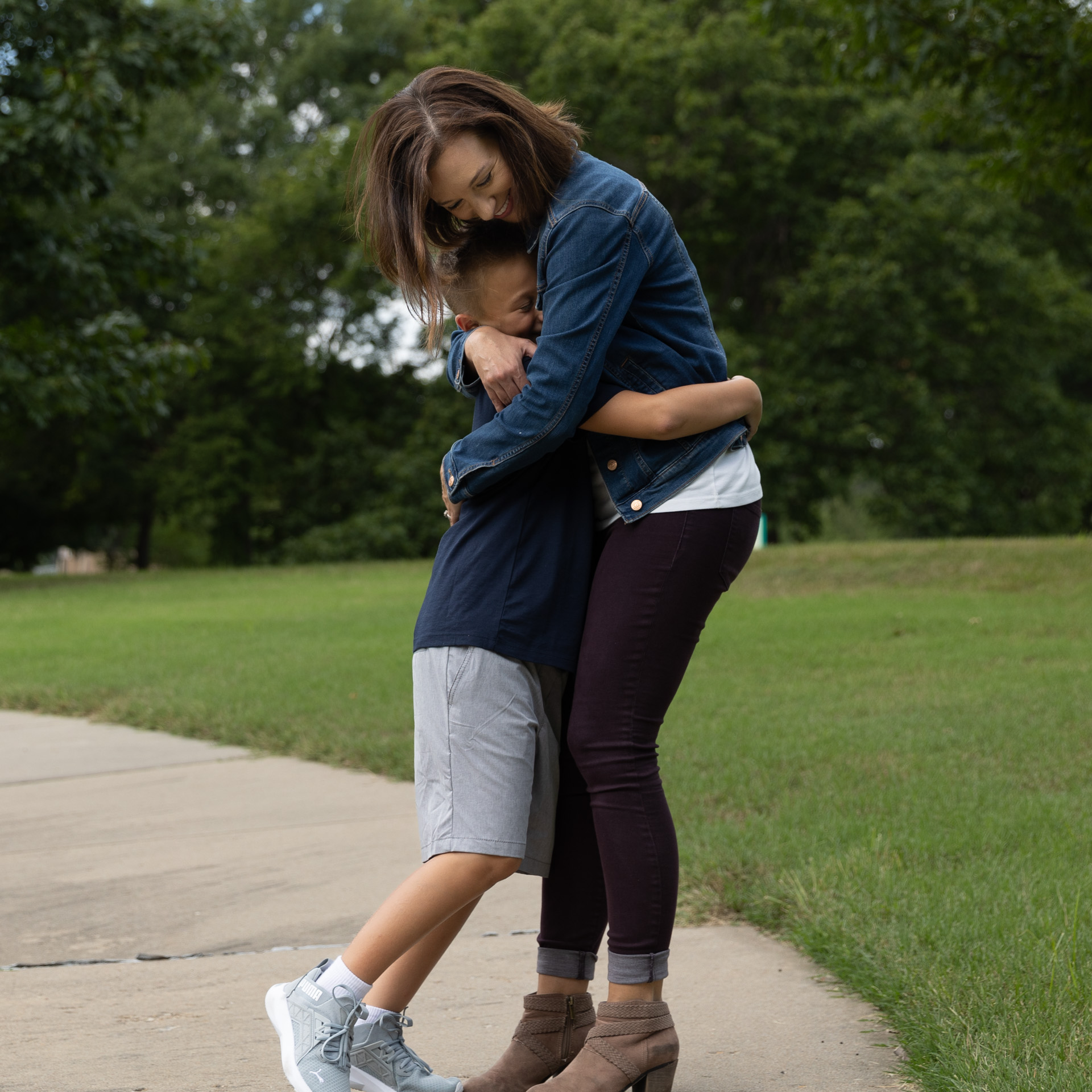 A tall dark-haired woman in a blue jean jacket warmly embraces a happy young boy who is wearing a blue T-Shirt and sneakers on a park path.