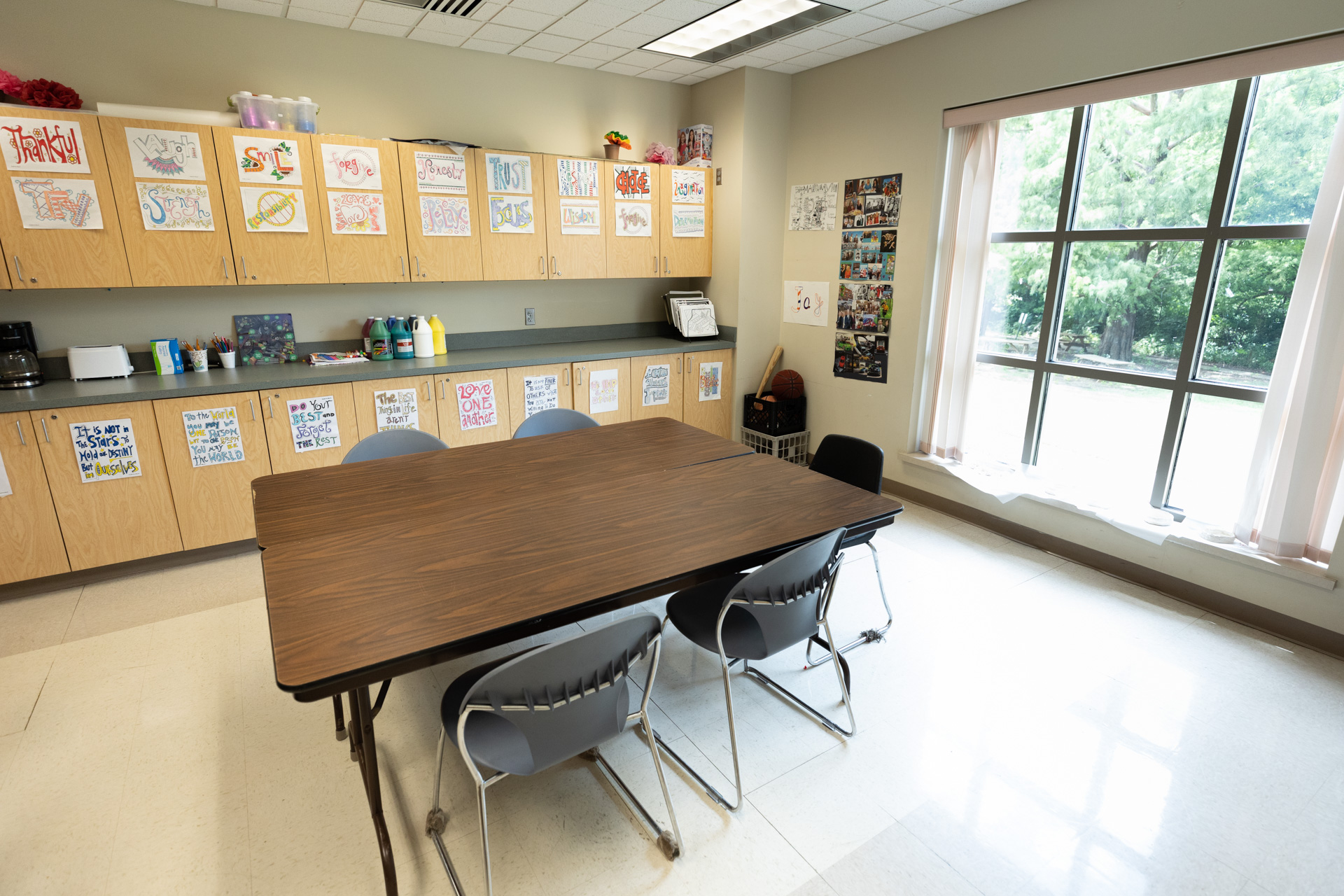 Inside classroom. Many cabinets at back of room with artwork. Large window on right side of room and image. Five-person table in center of room.
