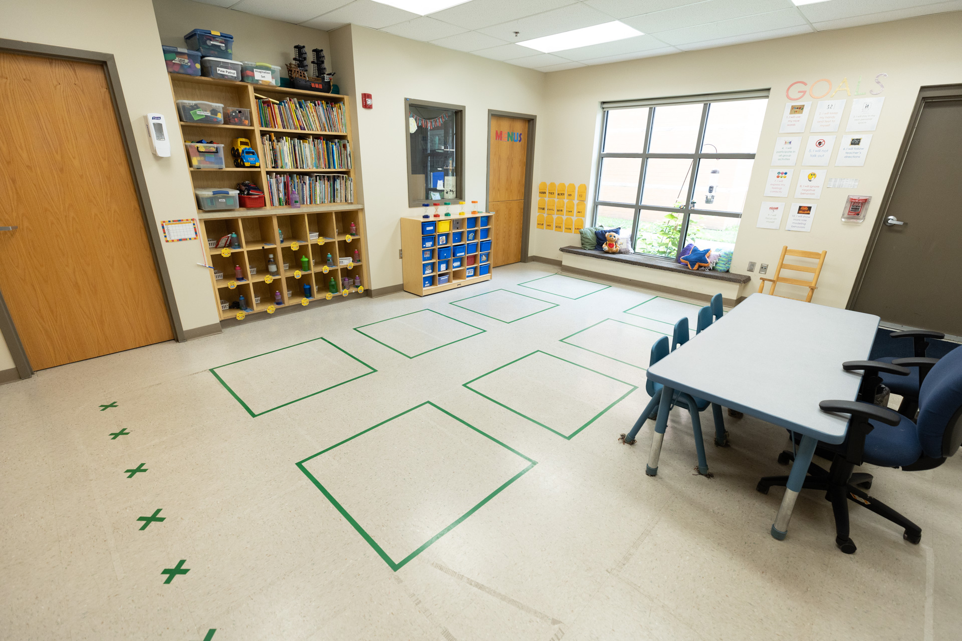 inside of classroom. Floor has eight squares taped on the ground. Many cubbies and cabinets along the back wall. Table with small chairs for children along the front wall.