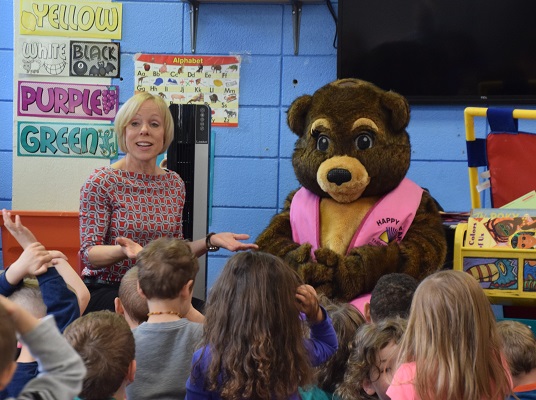 An FSGC Professional Stands At Front of Room With Bear Mascot (Person Inside of Bear Costume). Several Children Stand In Front of FSGC Professional and Bear, Facing Away From Camera. FSGC Professional Asks the Children a Question About the Bear with Arms Open and Palms Raised. Bear is Wearing Pink Stole With FSGC Logo.
