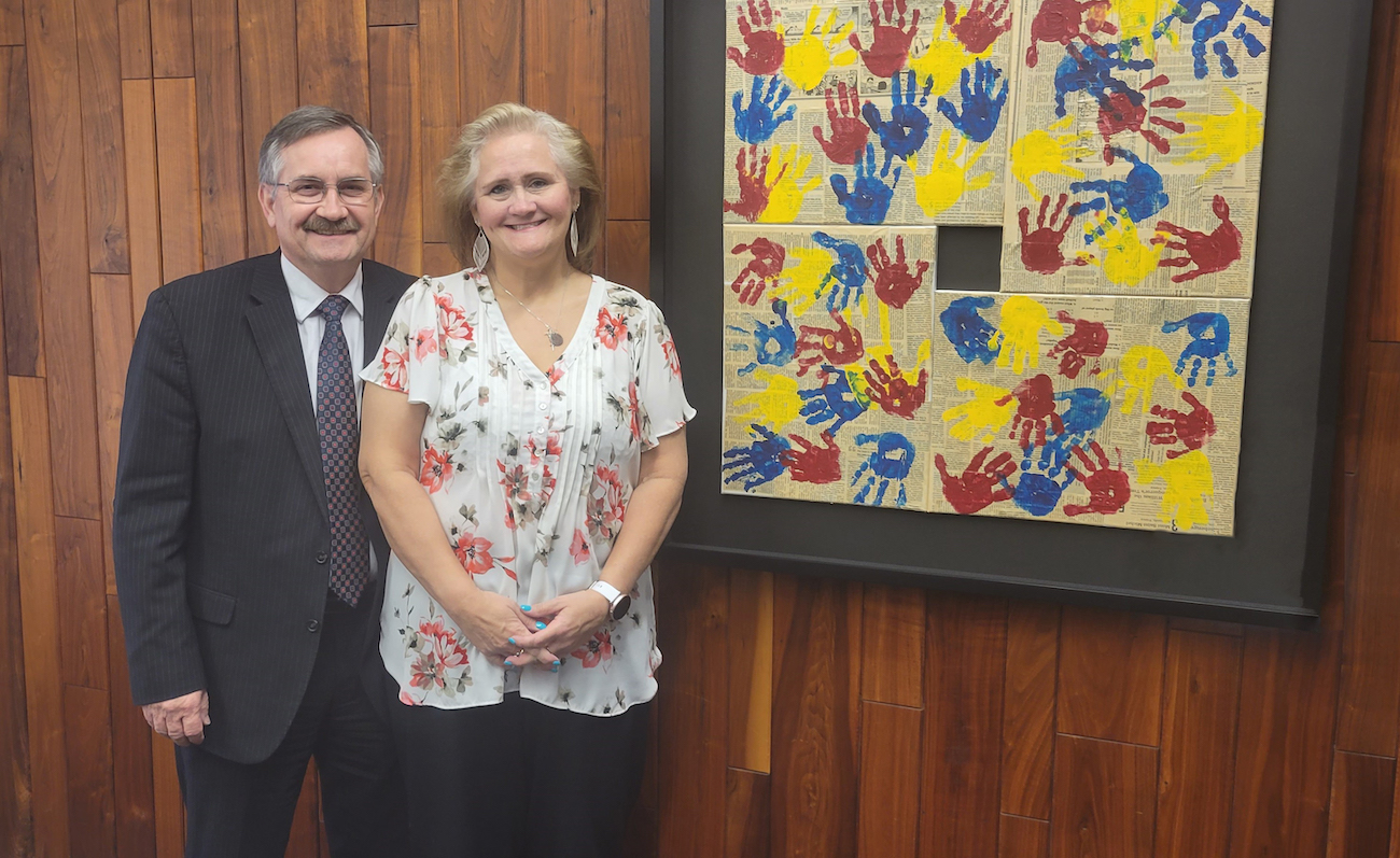 Two donors, Allan and Chris Towle, Stand In Front of Piece of Artwork. Art is Framed Square Shape with Blue, Red, and Yellow Handprints.
