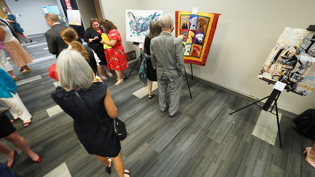 The silent auction room is filled with people looking at items displaced on tables. One of the items in a quilt with 9 dogs.