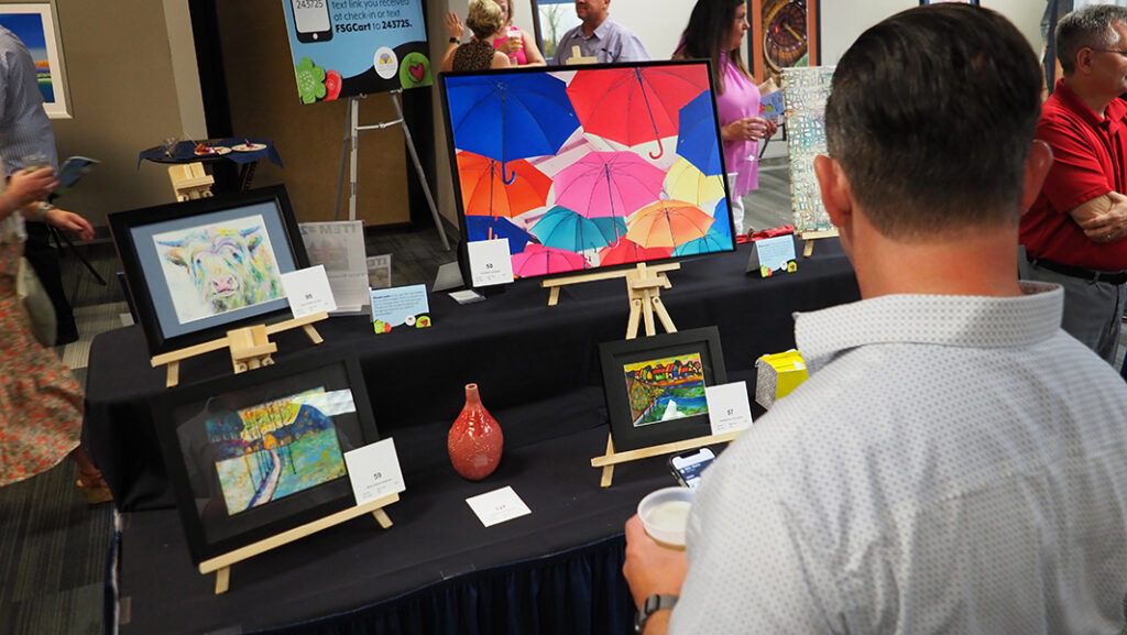 A guest looks at silent auctions items displayed on a table. Items include a pastel cow drawing, a colorful umbrella photograph, a fall painting of a house in Ukraine, a red ceramic vase, and a summer painting of a house in Ukraine.