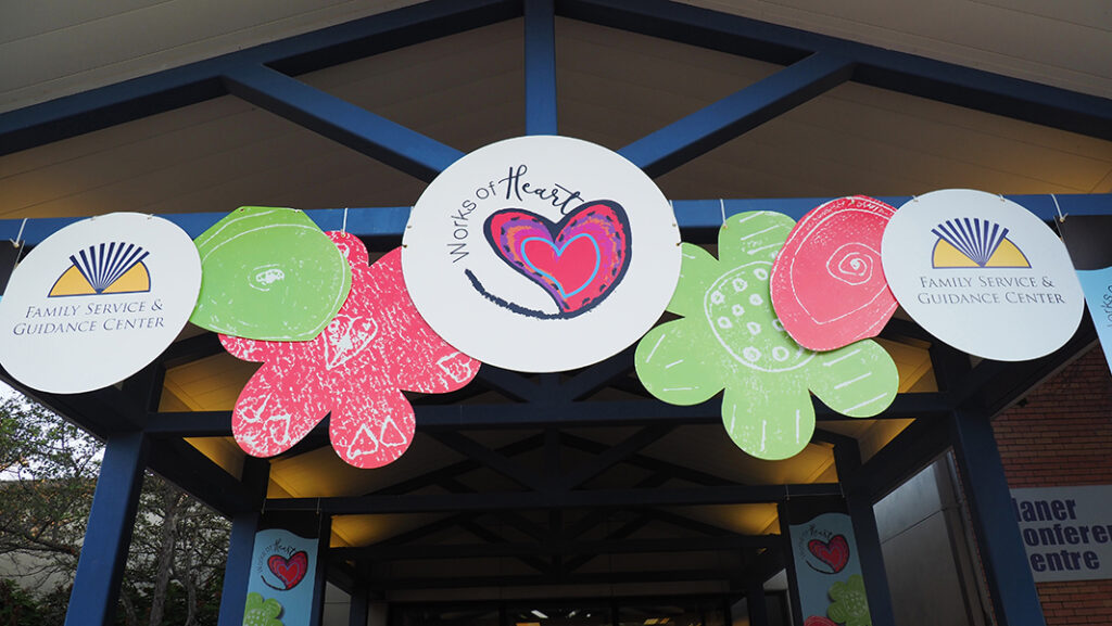 The entryway to the venue with large hanging signs. Two have the Family Service & Guidance Center logo, the other has the Works of Heart logo. Large graphic flowers are intermixed with the signs.