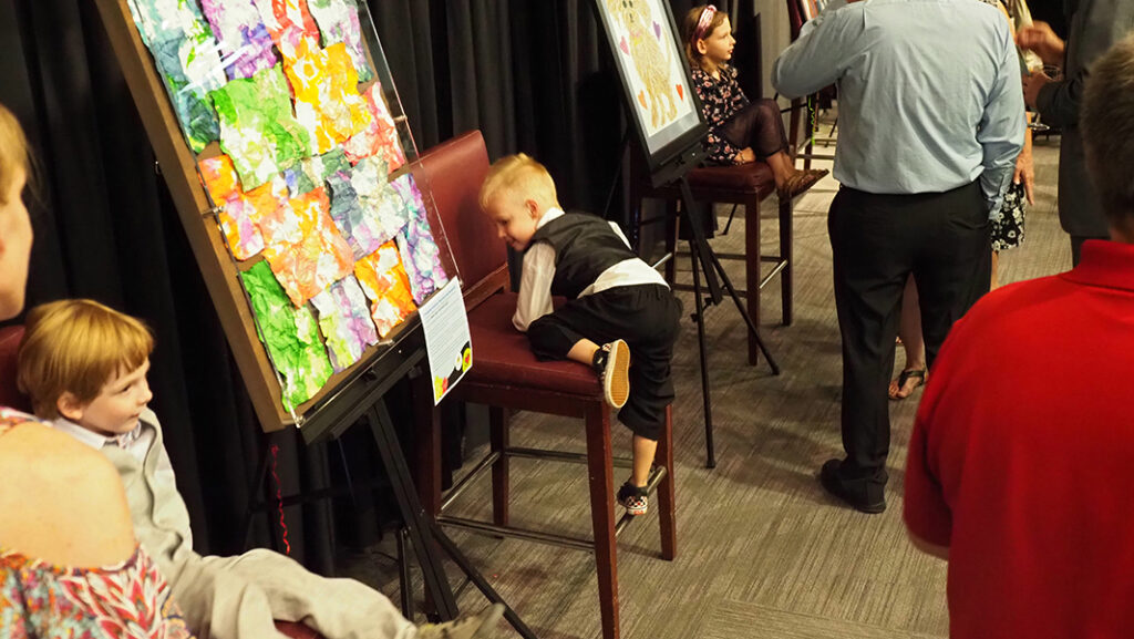 A male pre-school aged featured artist in a white shirt and black vest and pants crawls into his chair by his artwork. On the other side of the artwork is another male pre-school aged featured artist in a gray vest and pants. Their artwork is a collage of crumpled papers that have been dip-dyed green, orange, and purple.