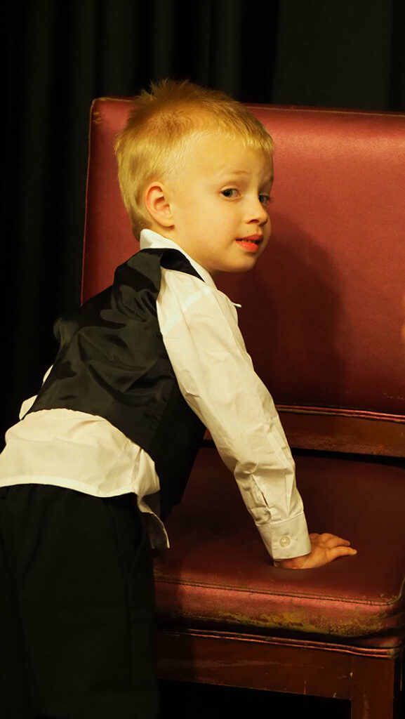 A male pre-school aged featured artist in a white shirt and black vest and pants crawls into his chair.