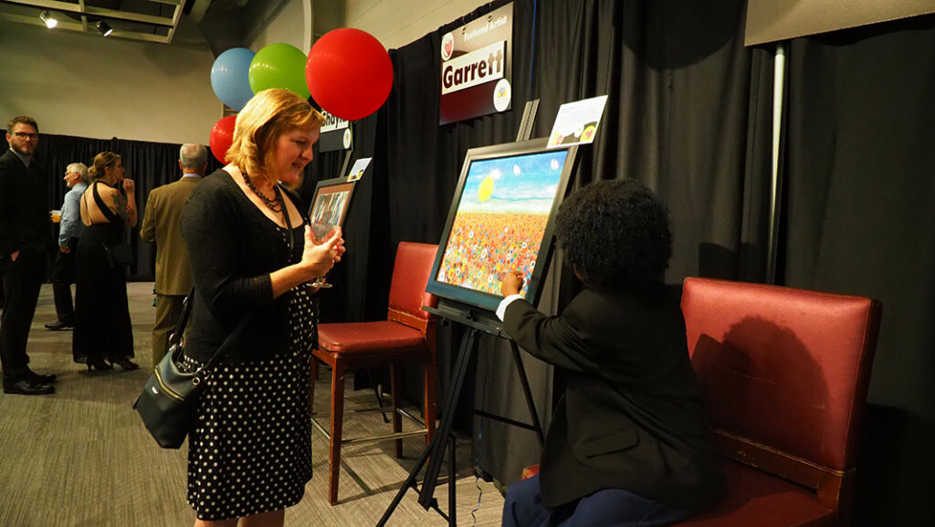 A young male featured artist with a black jacket sits in a chair and points to his artwork while talking to an adult female in a polka dotted dress. His artwork has a large yellow sun in a blue sky above a field of colorful flowers.