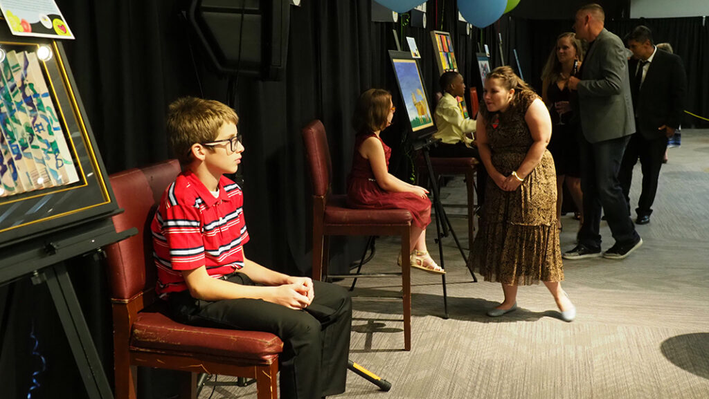 A young male featured artist with glasses and a striped polo shirt sits patiently in a chair by his artwork as adults guests make their way to talk to him.