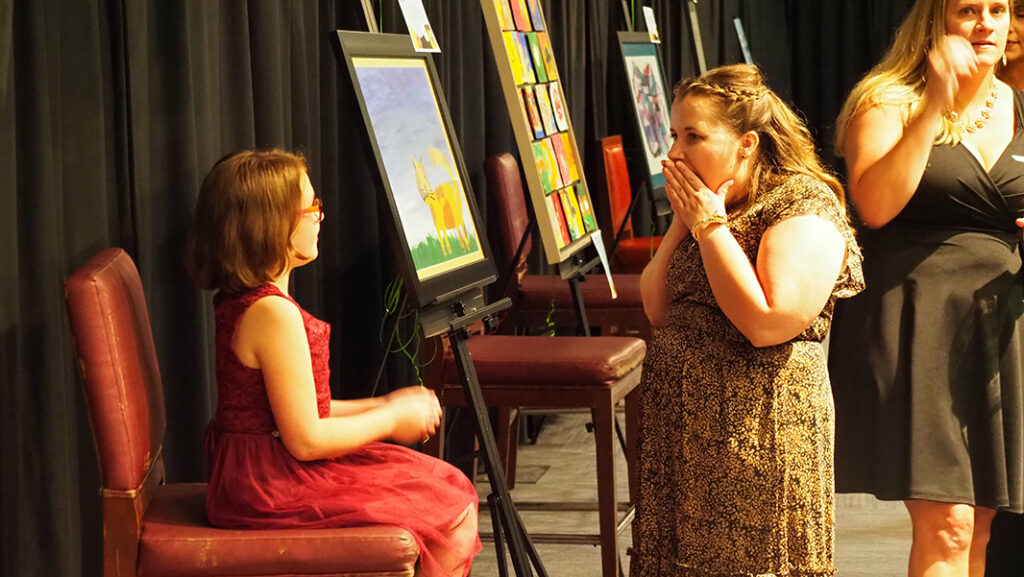 A young female featured artist in a maroon dress and glasses sits on a chair in front of her artwork and talks to an adult woman who is gasping with her hands over her mouth.