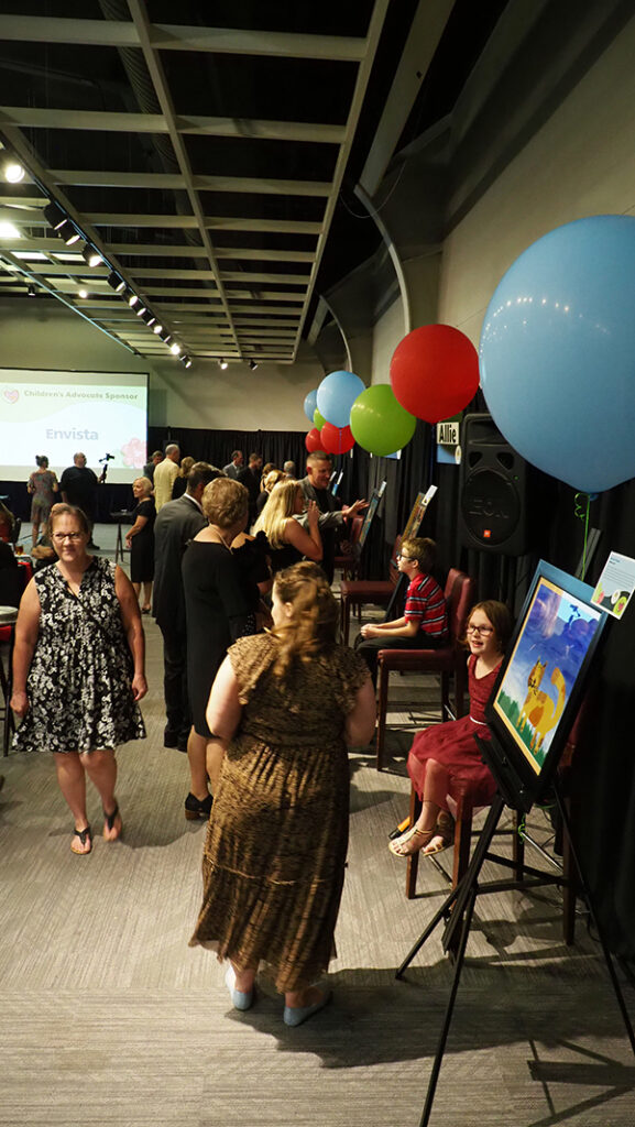 Guests walk around and talk to the client featured artists in chairs by their artwork. Large balloons are tied to each easel the artwork sits on.