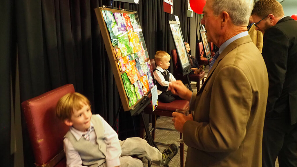 A pre-school aged male featured artist with a light purple shirt and gray vest and pants sits in a chair in front of his artwork, talking to a male in a brown suit.