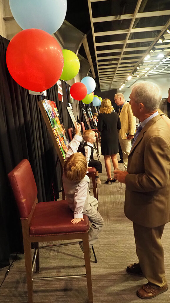A pre-school aged male featured artist with a light purple shirt and gray vest and pants points up to a large balloon while sitting in front of his artwork and talking to a male in a brown suit.