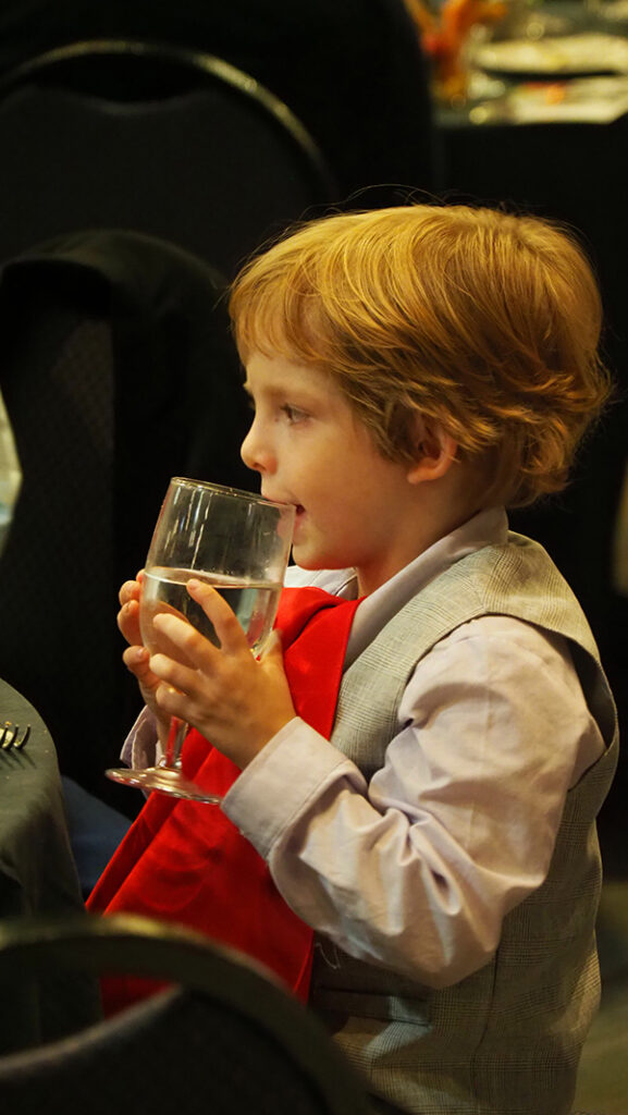 A pre-school aged male featured artists in a light purple shirt and gray vest takes a sip of water from a glass with a red napkin tucked into the neck of his shirt.