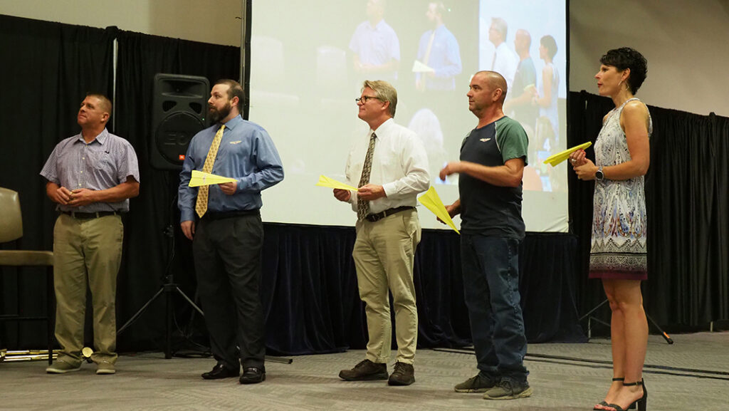 Five male and one female stand in line with their paper airplanes awaiting the final round on stage.