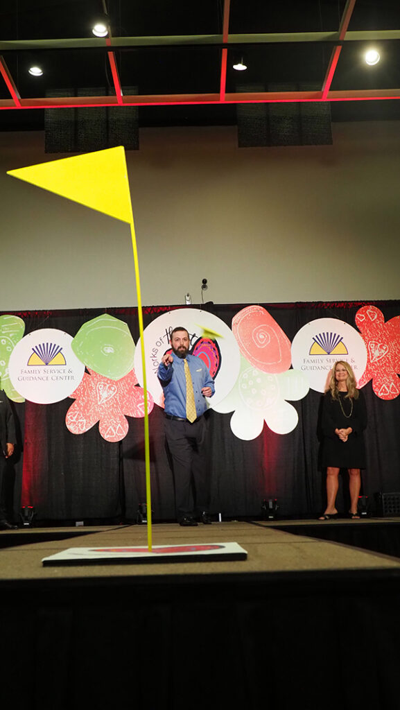 A neon flag is on stage as the target for the airplane toss finals. In the background is a male in a blue shirt and yellow tie throwing his paper airplane towards the target.