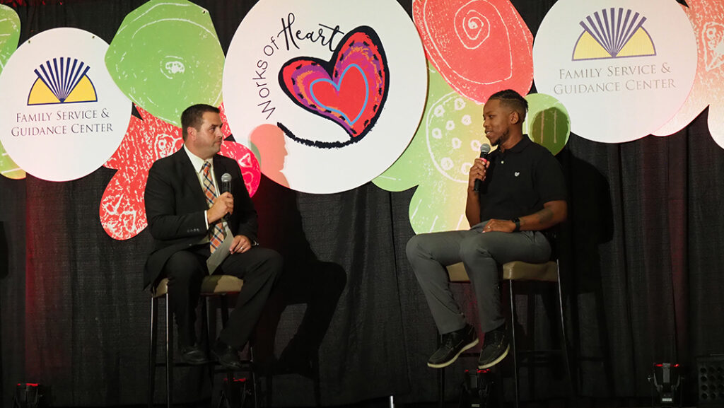 A male former client success story in a black polo shirt is interviewed on stage by a male in a suit.