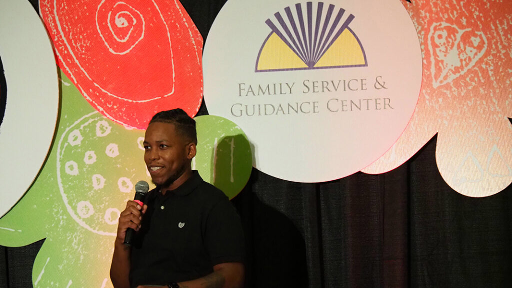A male former client success story in a black polo shirt speaks to the audience on stage with a microphone.