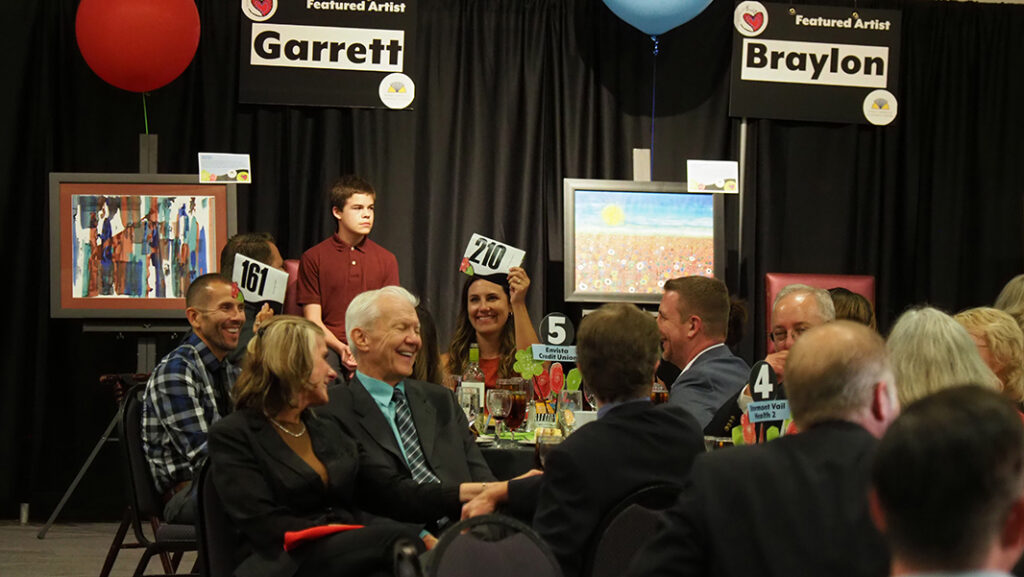 A male and female guest hold up their bidder numbers with smiles. A young male featured artist is in the background with a maroon shirt and his artwork.
