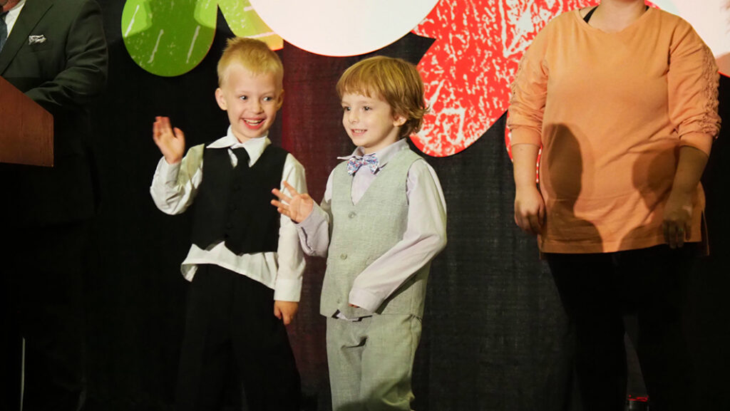 Two male pre-school aged featured artist, one in a white button up shirt with a black vest and pants, and the other with a light purple shirt and bowtie with a gray vest and pants, wave on stage while two male auctioneers speak to the crowd. 