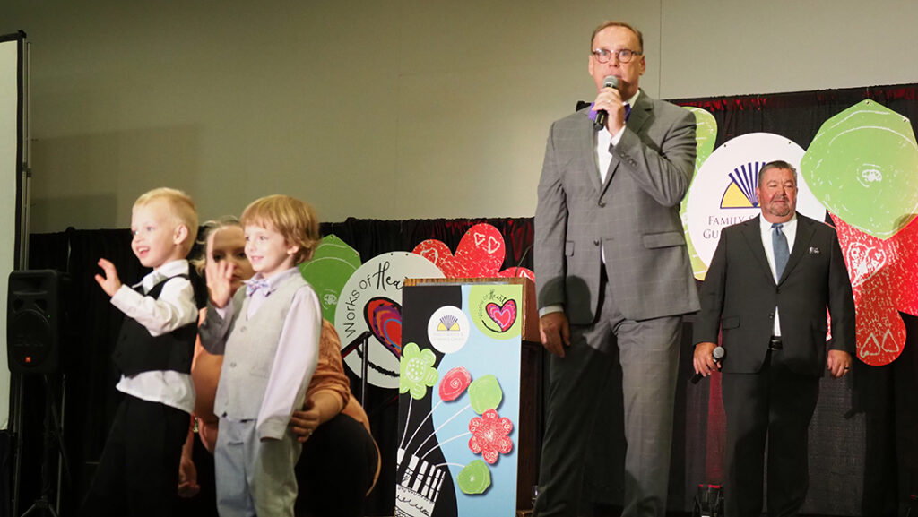 Two male pre-school aged featured artist, one in a white button up shirt with a black vest and pants, and the other with a light purple shirt and bowtie with a gray vest and pants, wave on stage while two male auctioneers speak to the crowd.