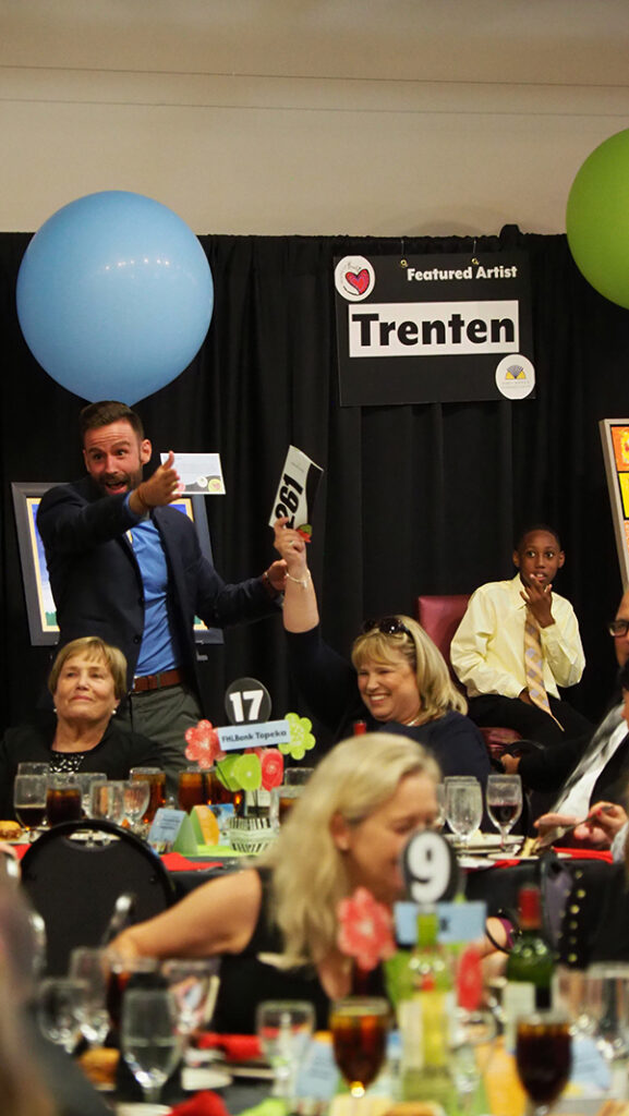An excited auction spotter points out a female bidder holding up her bidder number. Large colorful balloons are in the background and guests are sitting at tables.