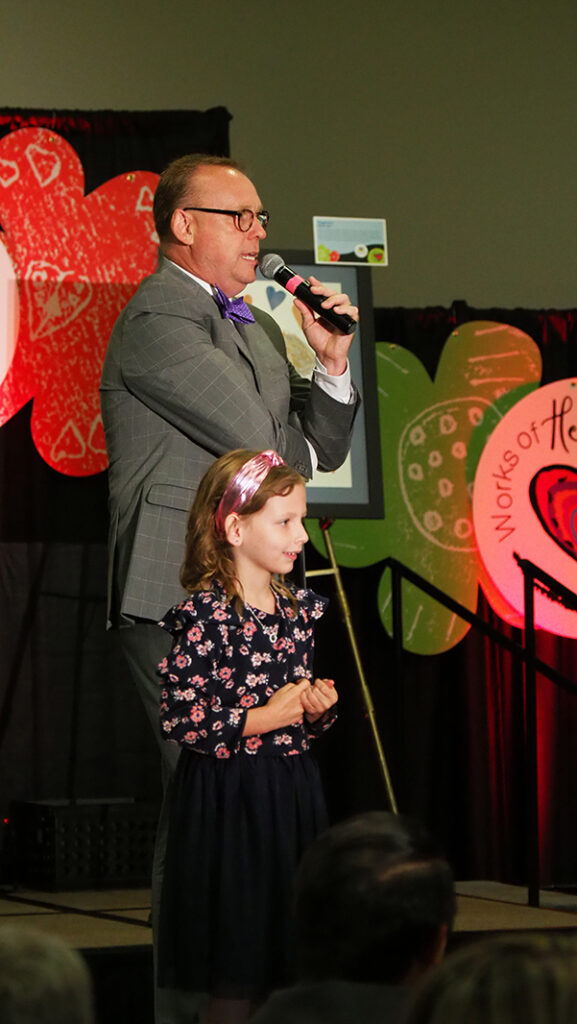 A male auctioneer speaking into a microphone on stage with a young female featured artist in a dress with a floral top and black bottom.
