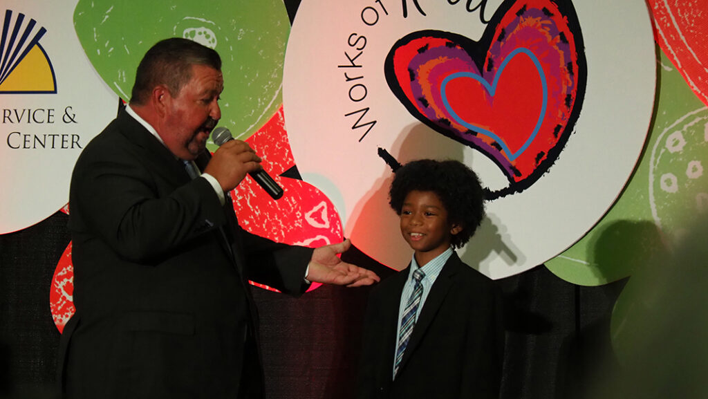 Male auctioneer with microphone talks to a young male smiling featured artist in a tie and jacket.