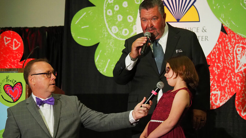 Two male auctioneers speaking to a young female featured artist in a maroon dress and glasses on stage.